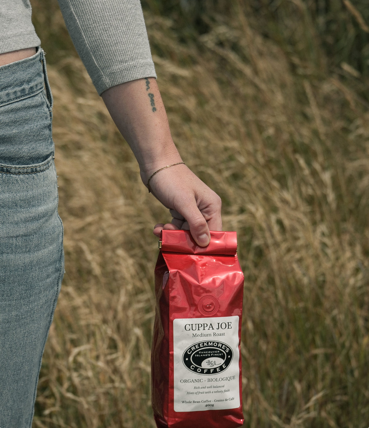 A person in a gray ribbed long-sleeve top and blue jeans holds a red bag of Cuppa Joe Greenhouse Coffee Roasted outdoors against a blurred field background.