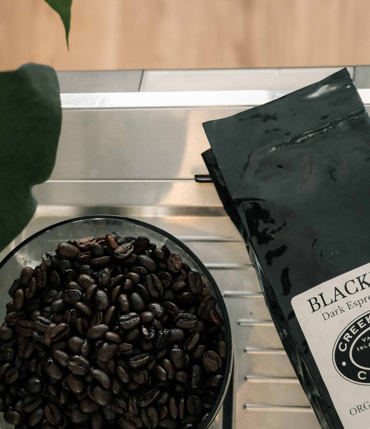 A black bag of Creek Coffee Co. Black Dark Espresso beans sits next to a glass bowl filled with roasted coffee beans on a stainless steel counter.