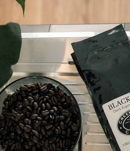 A black bag of Creek Coffee Co. Black Dark Espresso beans sits next to a glass bowl filled with roasted coffee beans on a stainless steel counter.