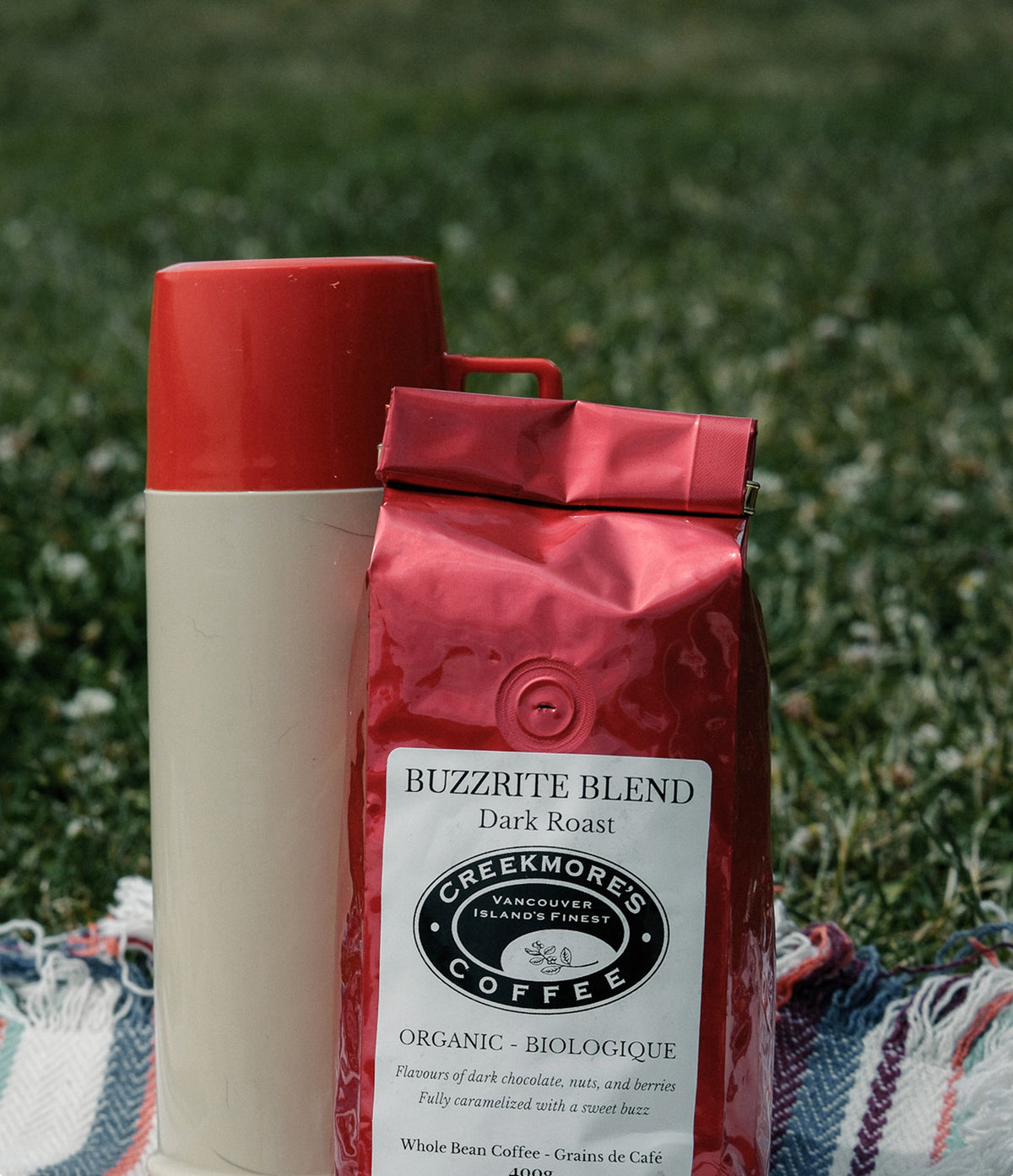 A bag of Creekmore's Coffee 'Buzzrite Blend' dark roast organic coffee stands next to a cream-colored bottle with a red cap on a red and white striped blanket, set against a blurred green grass background.