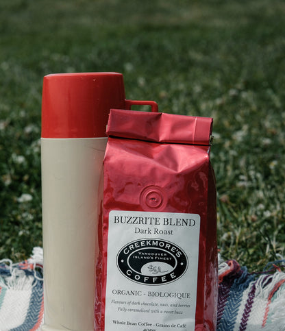 A bag of Creekmore's Coffee 'Buzzrite Blend' dark roast organic coffee stands next to a cream-colored bottle with a red cap on a red and white striped blanket, set against a blurred green grass background.