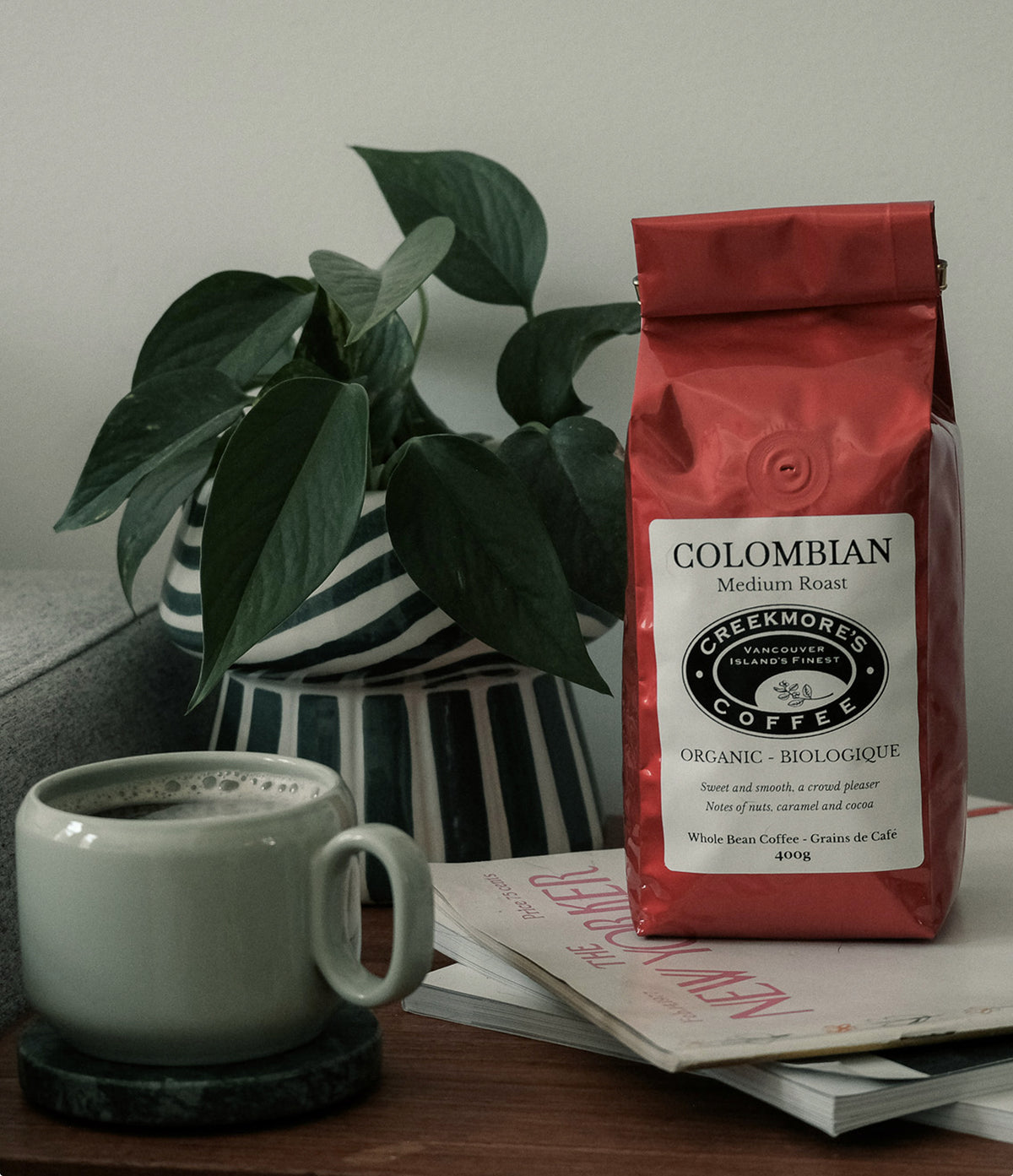 A red bag of Creekmore's Colombian Medium Roast coffee stands on a table next to a gray ceramic mug, a potted green plant, and a magazine.
