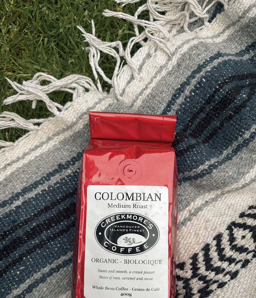 A red bag of Creekmore's Colombian Medium Roast organic coffee lies on a blue and white patterned woven blanket over green grass.