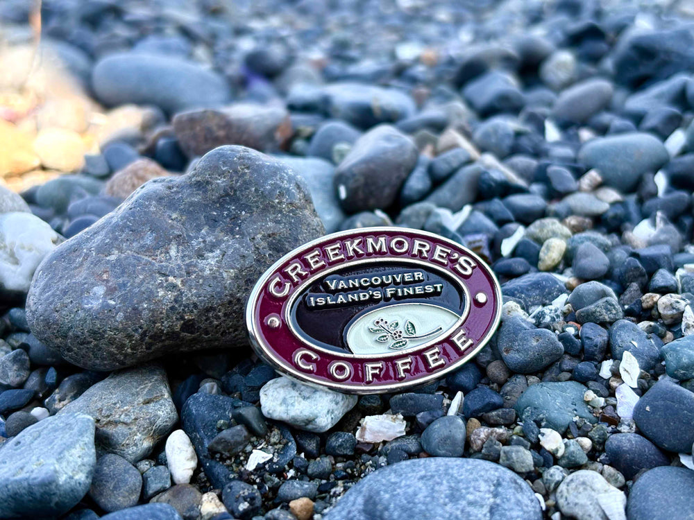 A circular enamel pin for Creekmore's Coffee, featuring a burgundy and silver design with the text 'Vancouver Island's Finest' and a coffee bean logo, resting on a bed of dark river pebbles.