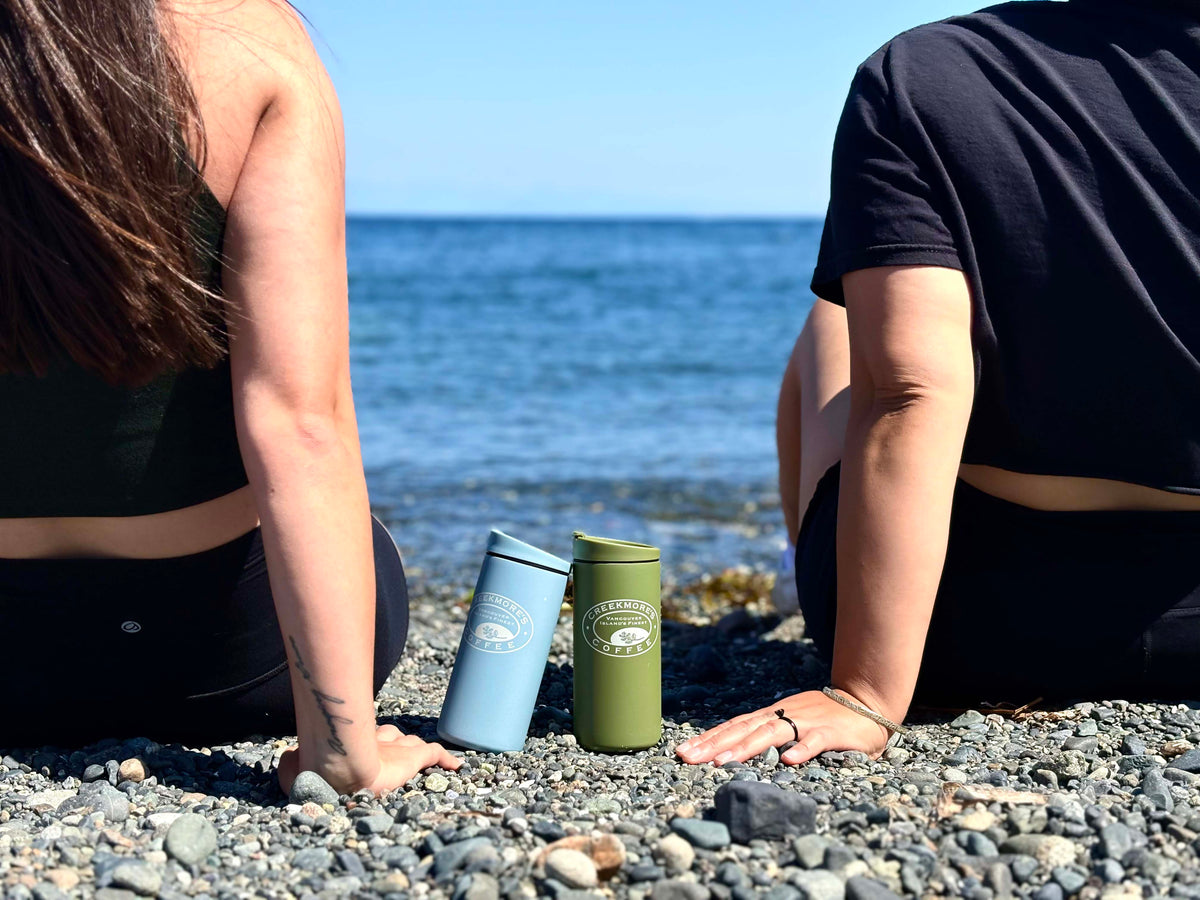 Two insulated travel tumblers, one light blue and one olive green, with a white 'The Coffee Club' logo, sitting on a pebble beach between two people looking out toward the ocean.
