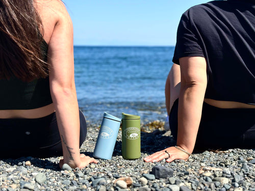Two insulated travel tumblers, one light blue and one olive green, with a white 'The Coffee Club' logo, sitting on a pebble beach between two people looking out toward the ocean.
