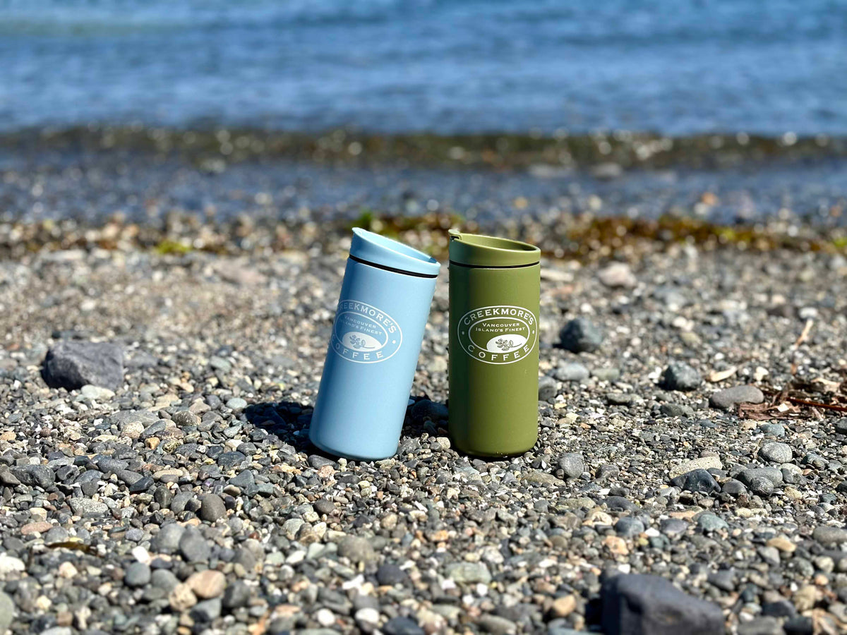 Two insulated travel tumblers standing on a pebble beach with water in the background. One tumbler is light blue and the other is olive green, both with white circular 'Pleasanton Coffee' logos on the front.