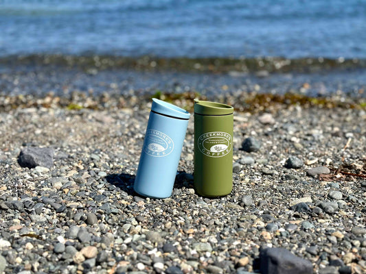 Two insulated travel tumblers standing on a pebble beach with water in the background. One tumbler is light blue and the other is olive green, both with white circular 'Pleasanton Coffee' logos on the front.