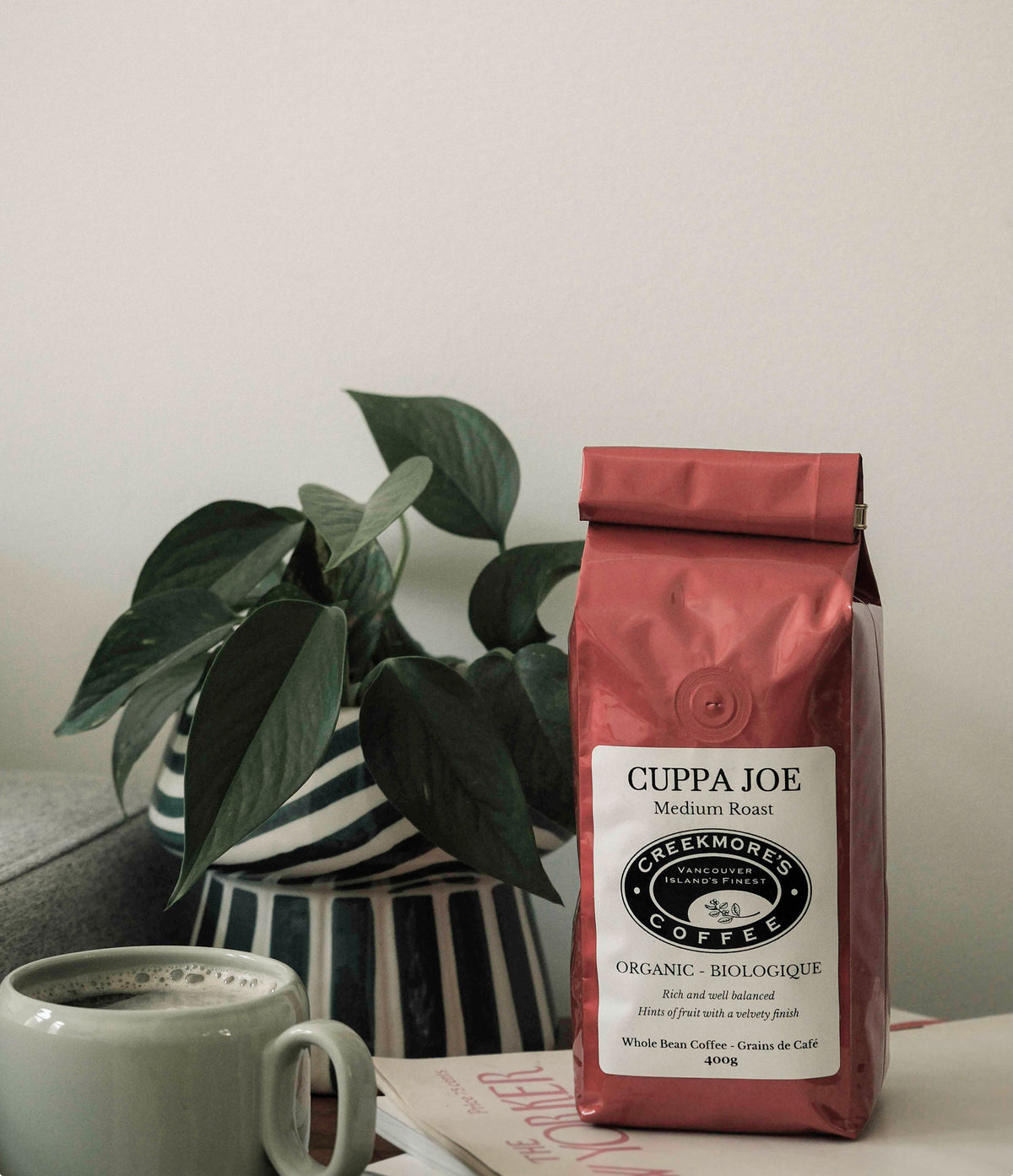 A bag of Cheekrohn's Coffee Cuppa Joe Medium Roast organic coffee stands on a tabletop next to a mug of coffee and a potted plant.