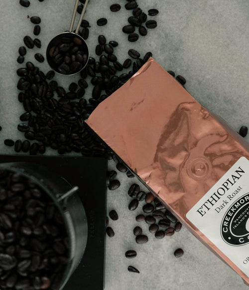 A bag of Ethiopian Dark Roast coffee from Creekstone Coffee Co. lies on a light gray surface surrounded by scattered roasted coffee beans, with a metal scoop filled with beans near the top left and a container of beans in the bottom left corner.