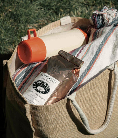 A beige canvas tote bag outdoors, filled with a striped knit blanket, an orange-capped insulated bottle, and a bag of Ethiopia-based coffee.