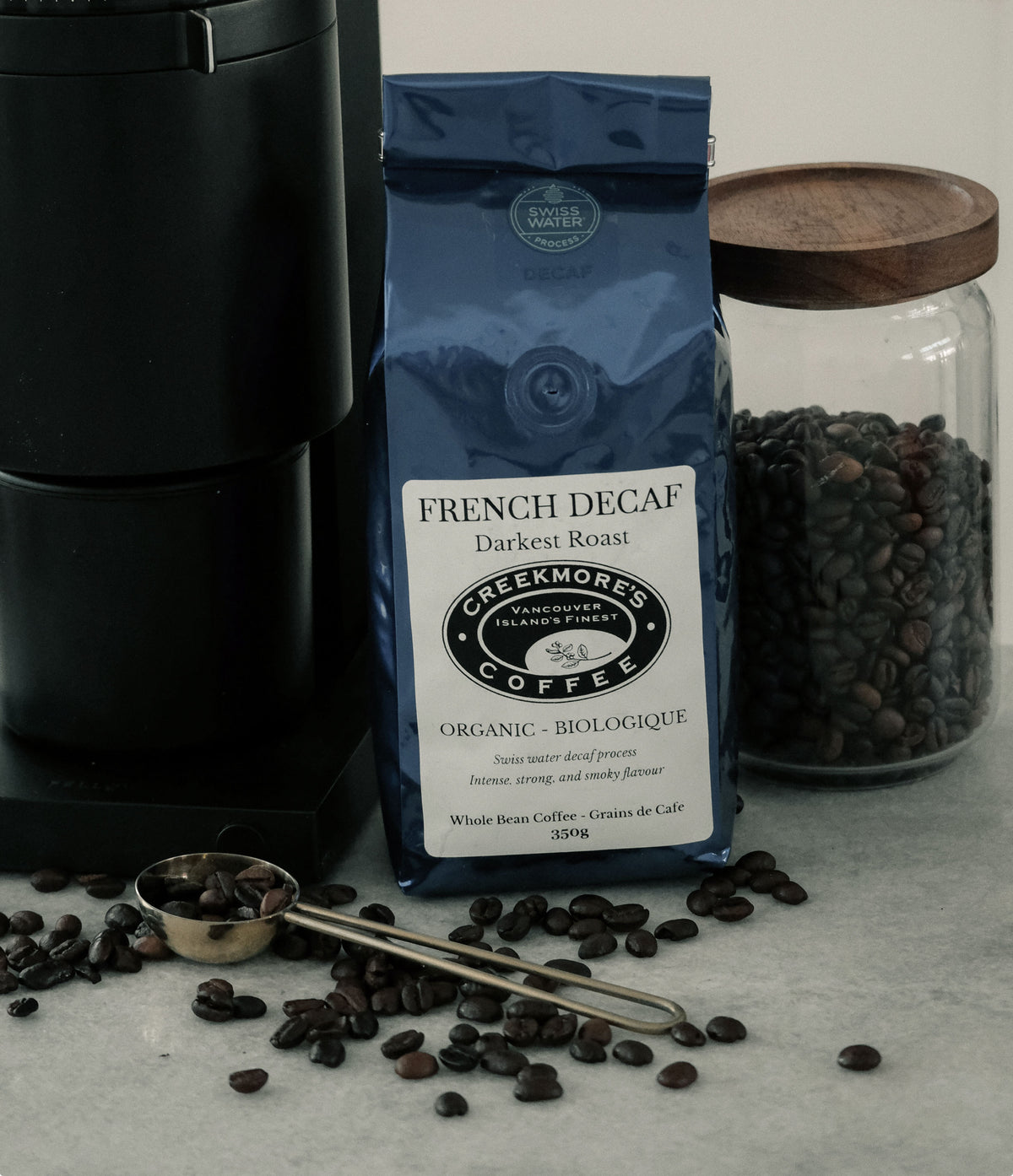 A bag of Creekmore's French Decaf Dark Roast coffee stands on a light marble surface, surrounded by scattered coffee beans, a metal measuring scoop, and a glass jar filled with beans. A black coffee maker is partially visible on the left.