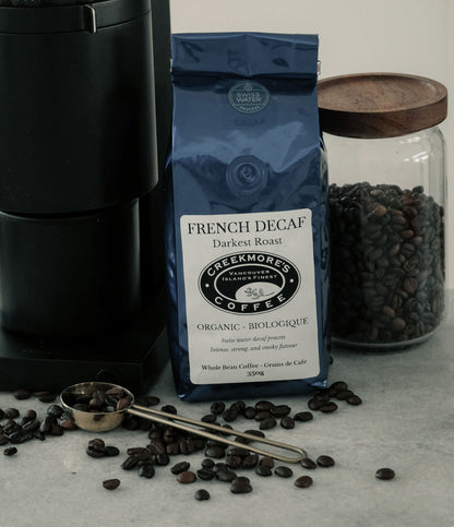 A bag of Creekmore's French Decaf Dark Roast coffee stands on a light marble surface, surrounded by scattered coffee beans, a metal measuring scoop, and a glass jar filled with beans. A black coffee maker is partially visible on the left.