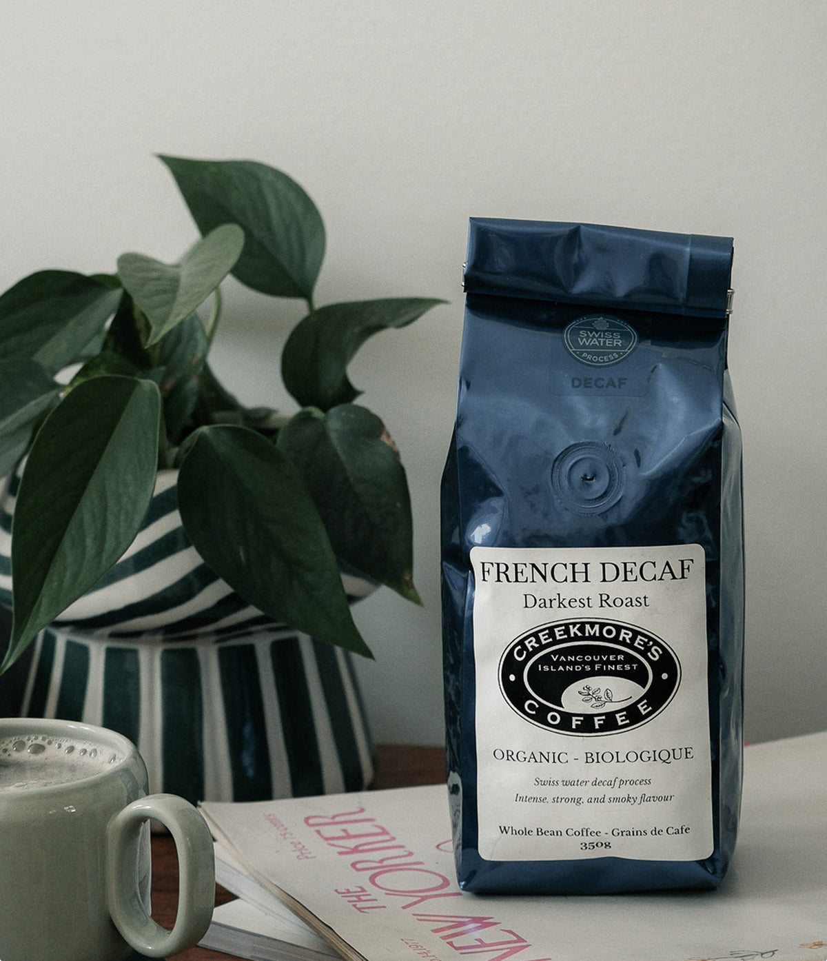 A bag of Creekmore's Coffee French Decaf Darkest Roast standing on a tabletop next to a green potted plant and a white mug, with a New Yorker magazine in front.