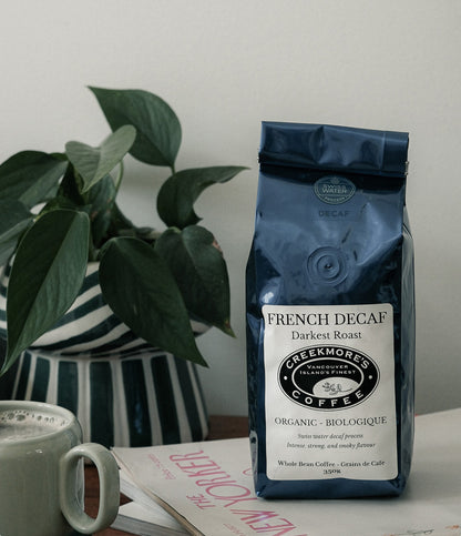 A bag of Creekmore's Coffee French Decaf Darkest Roast standing on a tabletop next to a green potted plant and a white mug, with a New Yorker magazine in front.