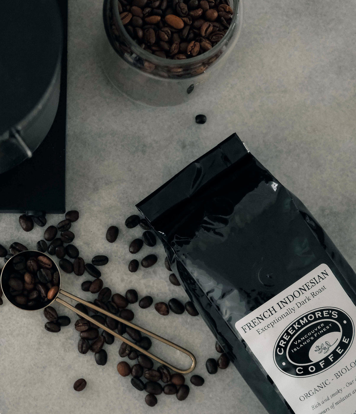 A bag of Greemore's French Indonesian coffee lies on a light stone surface with scattered coffee beans, a metal scoop filled with beans, and a glass jar of roasted beans nearby.