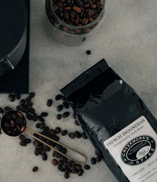 A bag of Greemore's French Indonesian coffee lies on a light stone surface with scattered coffee beans, a metal scoop filled with beans, and a glass jar of roasted beans nearby.