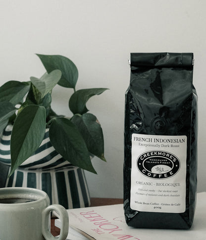 A bag of Creekmonos Coffee French Indonesian V60 Organic Dark Roast whole bean coffee stands on a table next to a white mug and a potted green plant against a plain white wall.