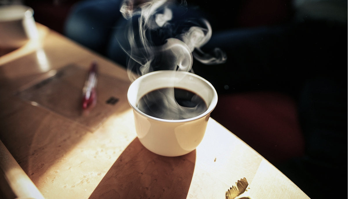 A white ceramic cup filled with black coffee, with visible steam rising, set on a wooden table in warm, low light.