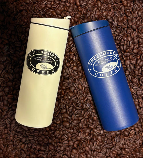 Two insulated travel tumbler cups from Creekmore's Coffee, one cream and one blue, displayed on a bed of roasted coffee beans. The cream cup has a black lid and black logo, while the blue cup has a white logo.