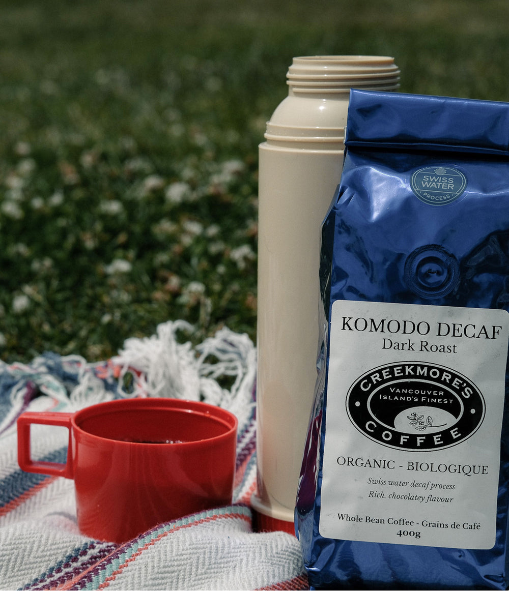 A bag of Creekmore's Coffee Komodo Decaf Dark Roast stands next to a cream-colored vacuum flask and a red ceramic mug on a patterned blanket outdoors on a grassy lawn.