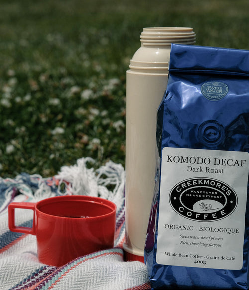 A bag of Creekmore's Coffee Komodo Decaf Dark Roast stands next to a cream-colored vacuum flask and a red ceramic mug on a patterned blanket outdoors on a grassy lawn.