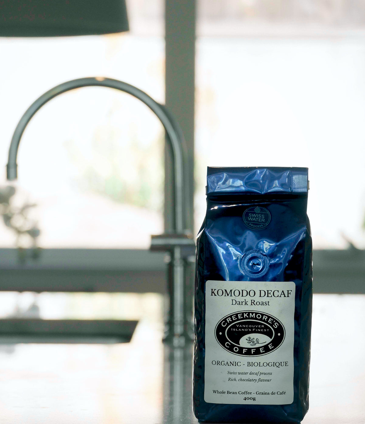 A bag of Creekmore's Coffee Komodo Decaf Dark Roast standing on a kitchen counter in front of a blurred faucet and bright window.