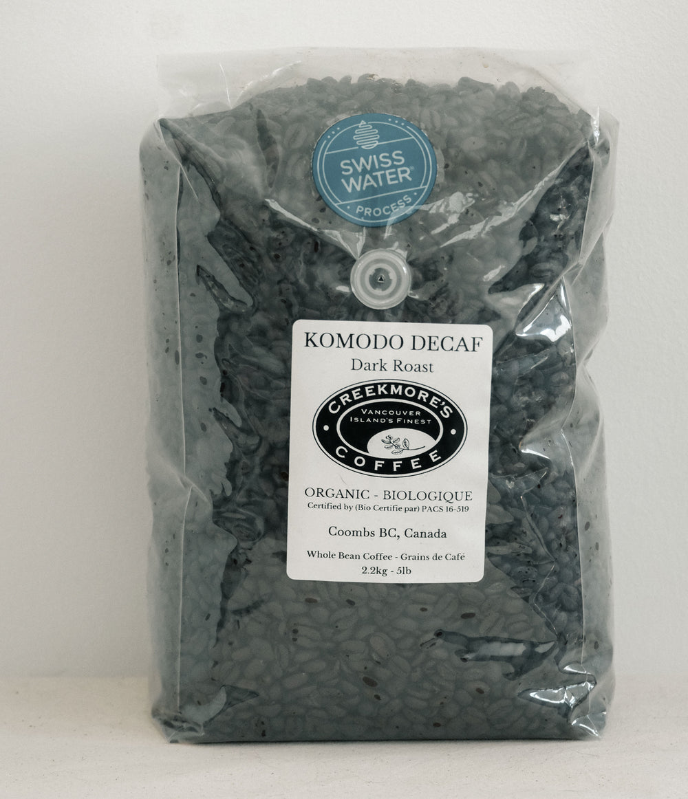 A bag of Creekmore's Coffee Komodo Decaf Dark Roast organic whole bean coffee, sealed in a clear plastic bag with a white label and a blue 'Swiss Water Process' sticker.