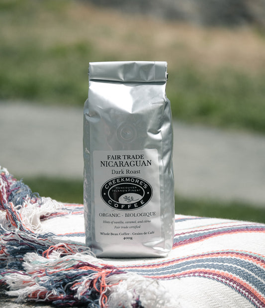 A silver bag of Greekmores Coffee Fair Trade Nicaragua Dark Roast stands on a striped woven blanket outdoors with a blurred natural background.