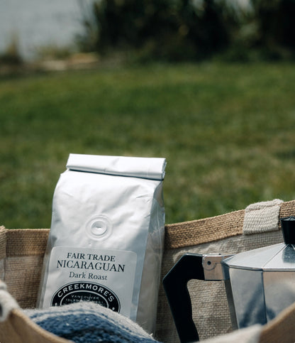 A bag of Creekmore's Fair Trade Nicaraguan Dark Roast coffee sits in a burlap basket next to a stovetop moka pot, with a blurred green grassy background.