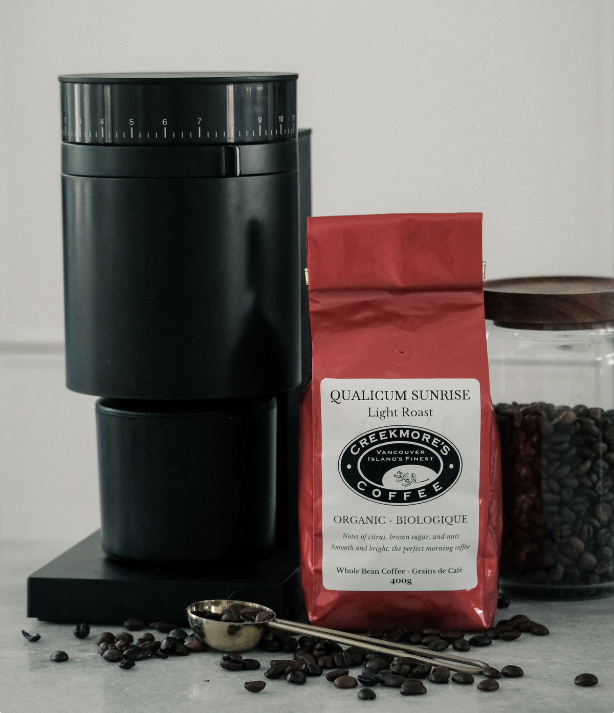 A black manual coffee grinder stands next to a red bag of Creekmore's Qualicum Sunrise Light Roast organic coffee, with scattered coffee beans on a light gray surface and a glass jar of coffee beans in the background.