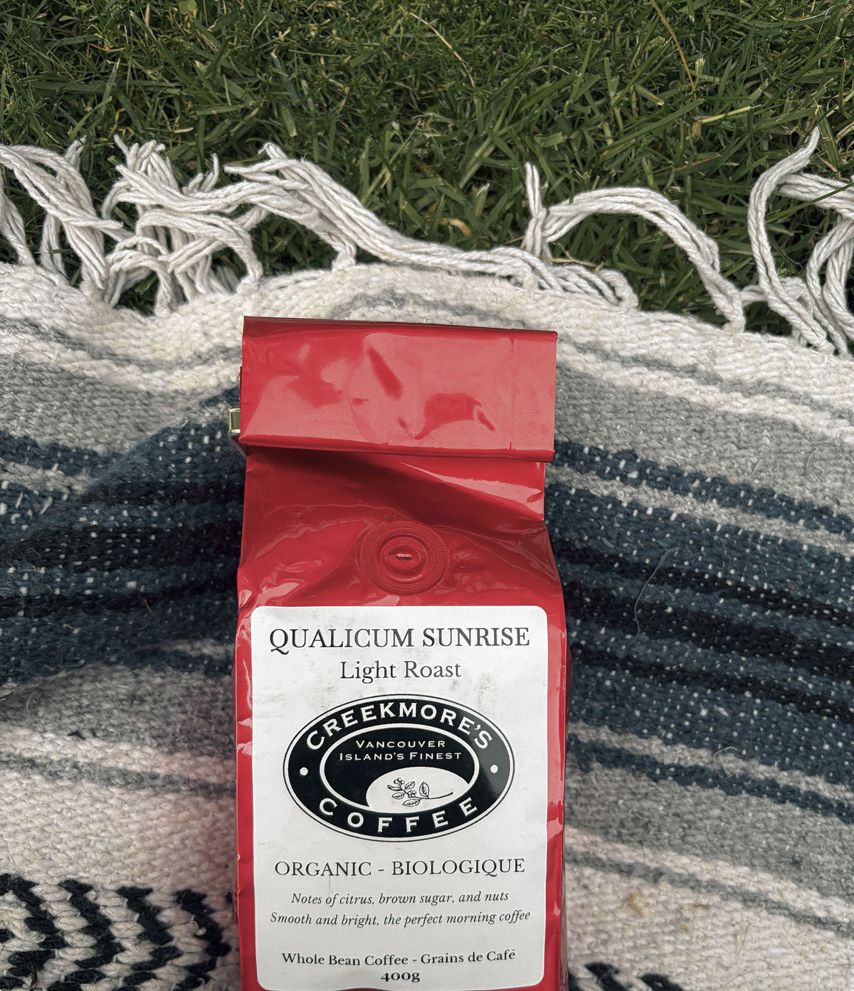 A red bag of Creekmore Coffee Qualicum Sunrise Light Roast whole bean coffee, placed on a blue and white striped woven blanket with green grass in the background.