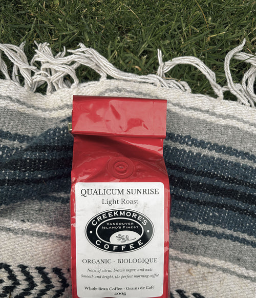 A red bag of Creekmore Coffee Qualicum Sunrise Light Roast whole bean coffee, placed on a blue and white striped woven blanket with green grass in the background.