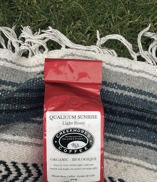 A red bag of Creekmore Coffee Qualicum Sunrise Light Roast whole bean coffee, placed on a blue and white striped woven blanket with green grass in the background.