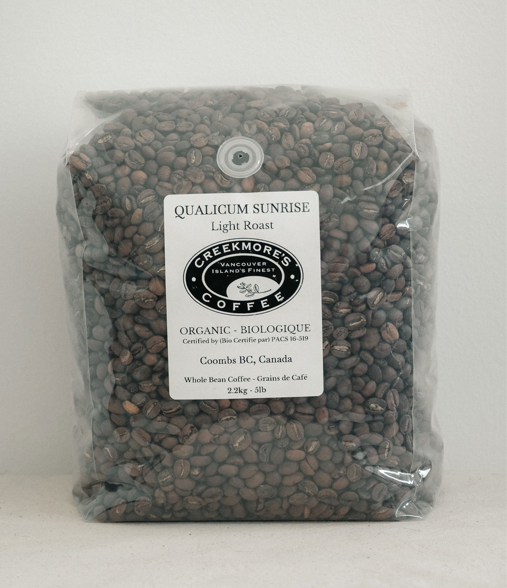 A clear plastic bag filled with dark brown coffee beans, featuring a white label for Creekmore's Coffee, Qualicum Sunrise Light Roast, Coombs BG, Canada.