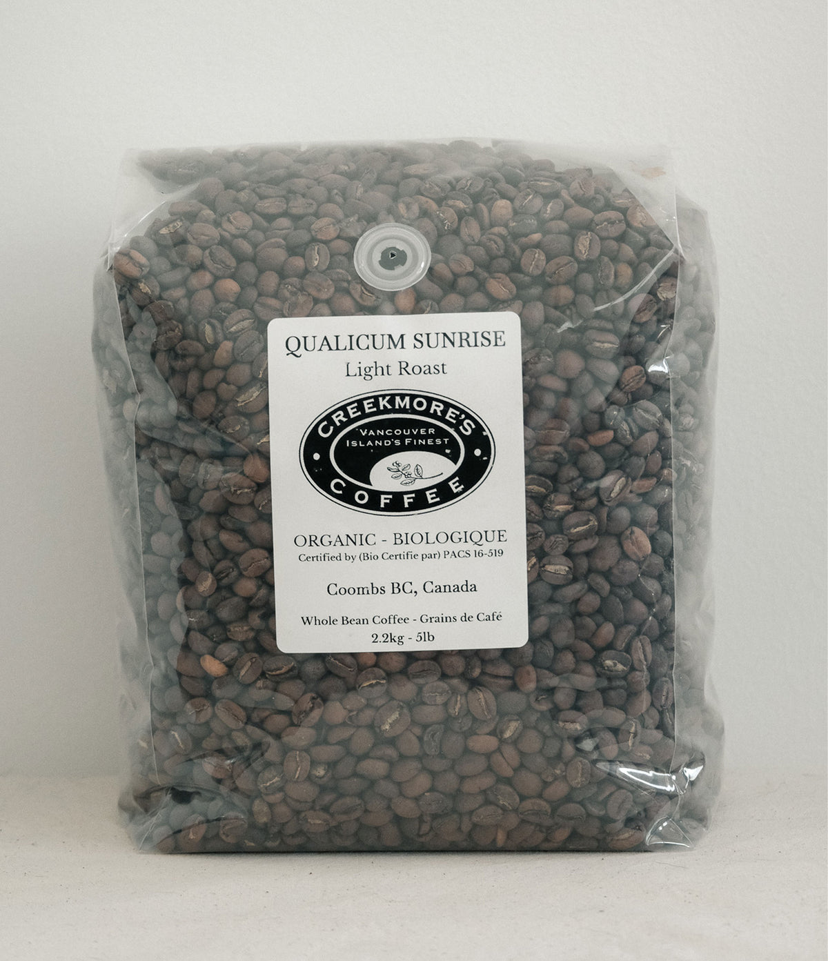 A clear plastic bag filled with dark brown coffee beans, featuring a white label for Creekmore's Coffee, Qualicum Sunrise Light Roast, Coombs BG, Canada.