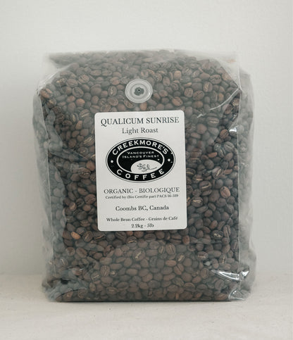 A clear plastic bag filled with dark brown coffee beans, featuring a white label for Creekmore's Coffee, Qualicum Sunrise Light Roast, Coombs BG, Canada.