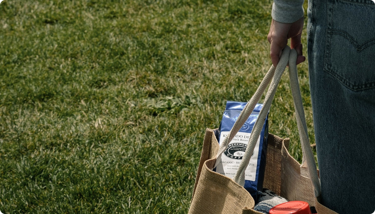 A person wearing blue jeans holds a tan burlap grocery tote bag with white handles on a green grass lawn. Inside the bag are packaged items, including a blue bag with white text.