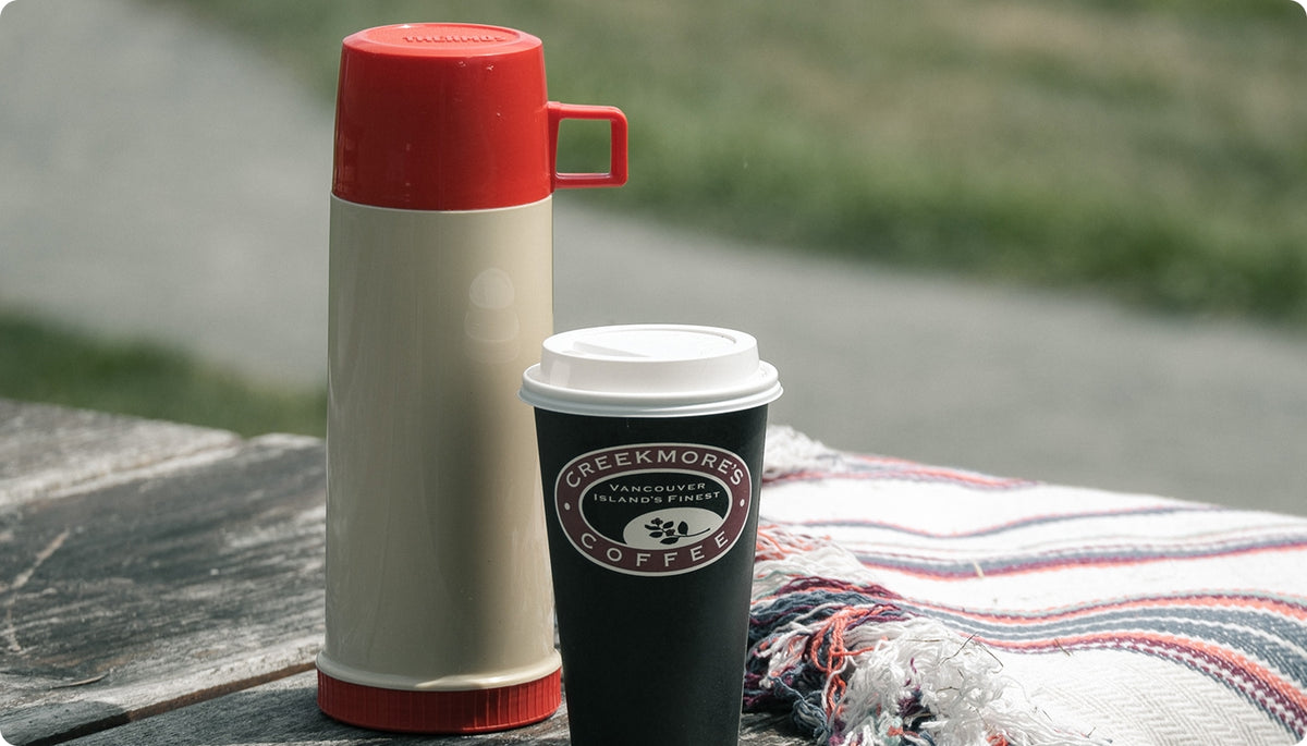 A vintage-style cream and red thermos bottle stands next to a black paper coffee cup with a white lid on a rustic wooden table, with a striped blanket and a blurred outdoor background.