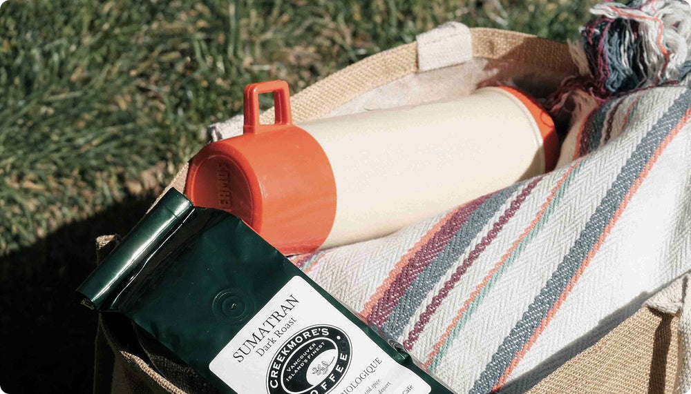A styled outdoor picnic scene featuring a cream and orange insulated bottle, a striped woven blanket, and a bag of Sumitran Greenbournes Coffee on a burlap picnic basket with grass in the background.