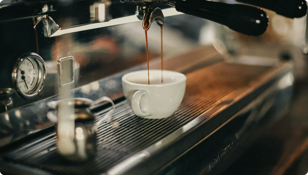 A close-up shot of espresso dripping from an espresso machine into a small white ceramic cup on a metal drip tray.