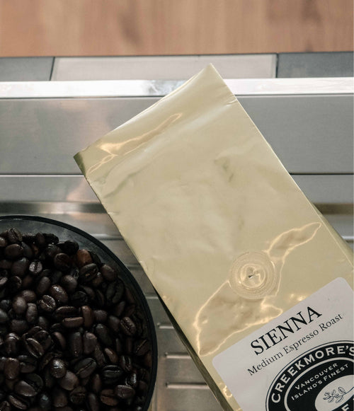 A bag of Creekmore's Sienna Medium Espresso Roast coffee beans resting on a stainless steel counter next to a black bowl filled with roasted coffee beans.