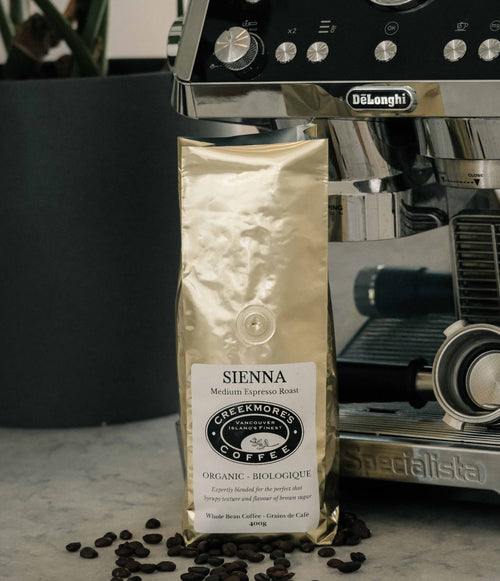 A bag of Creekmore Coffee Sienna Medium Roast coffee beans stands on a marble surface with scattered coffee beans in front and a De'Longhi espresso machine in the background.