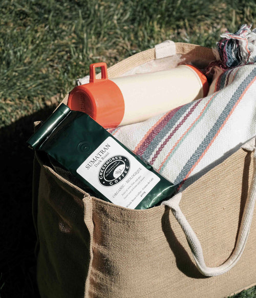 A beige jute tote bag outdoors on grass, filled with a striped blanket, a cream bottle with an orange cap, and a dark green coffee bag labeled Sumaytan Coffee.