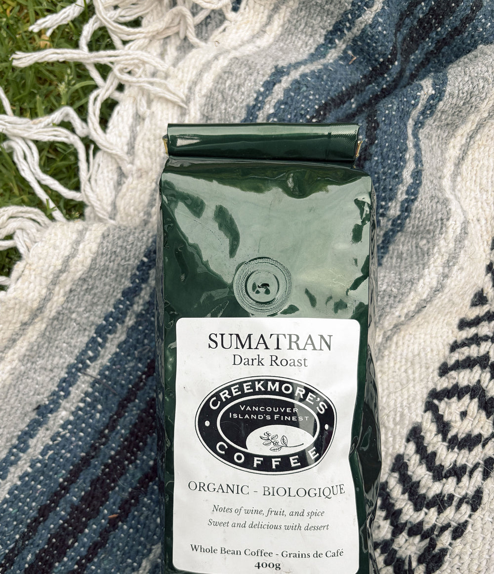 A 400g bag of Creekmore's Sumatran Dark Roast organic coffee, placed on a blue and white patterned knit blanket with green grass visible in the background.