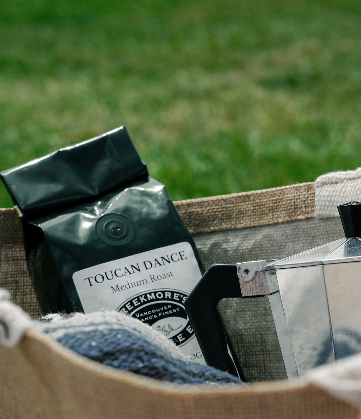 A bag of Toucan Dance Medium Roast coffee sits in a woven burlap basket outdoors, with a metal moka pot and blue fabric visible against a blurred green grass background.