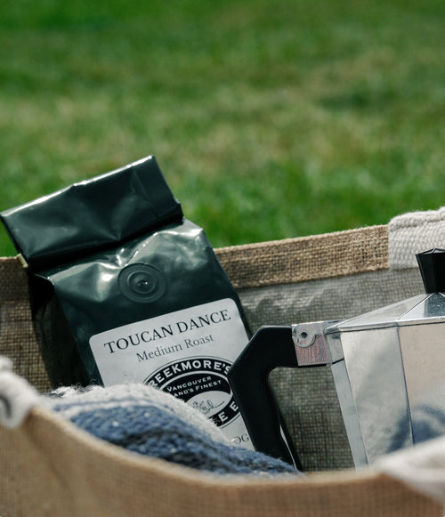 A bag of Toucan Dance Medium Roast coffee sits in a woven burlap basket outdoors, with a metal moka pot and blue fabric visible against a blurred green grass background.