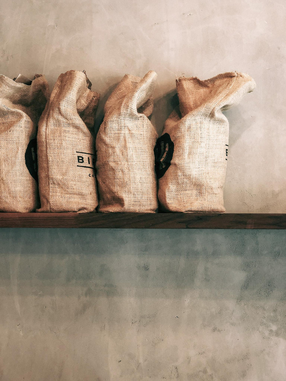 Four natural burlap coffee bags standing upright on a dark wooden shelf against a textured concrete wall.