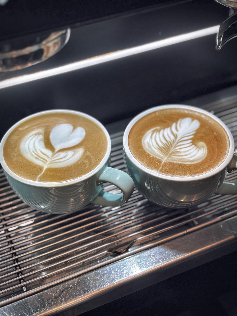 Two ceramic coffee cups filled with latte art sit on a stainless steel espresso machine drip tray. The cups are a pale blue-green with ribbed texture, and the coffee has white leaf and heart-shaped latte designs on top.