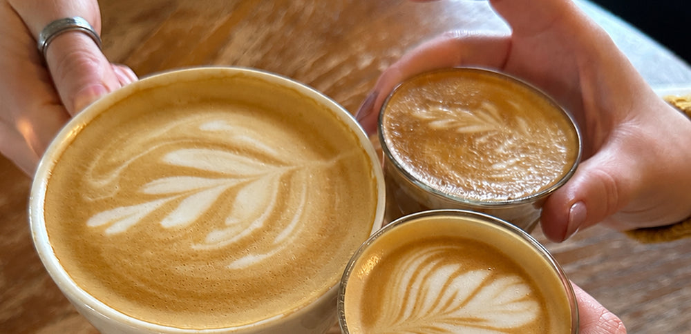 Three cups of latte coffee with leaf latte art, including one large ceramic cup and two small metal travel cups, held by hands over a wooden table.